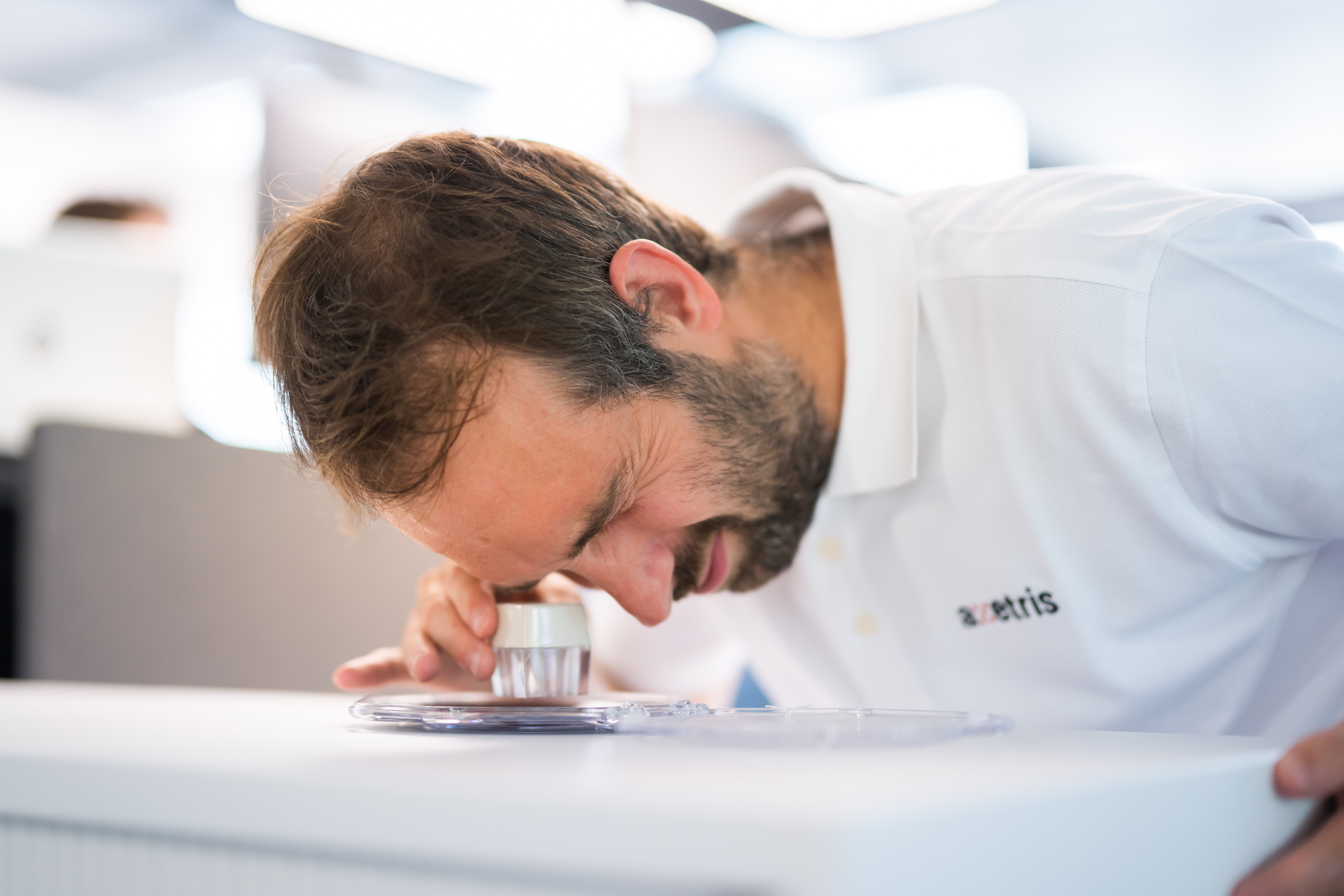 A person examines a sample in a laboratory setting, closely observing a petri dish while wearing a white lab coat.