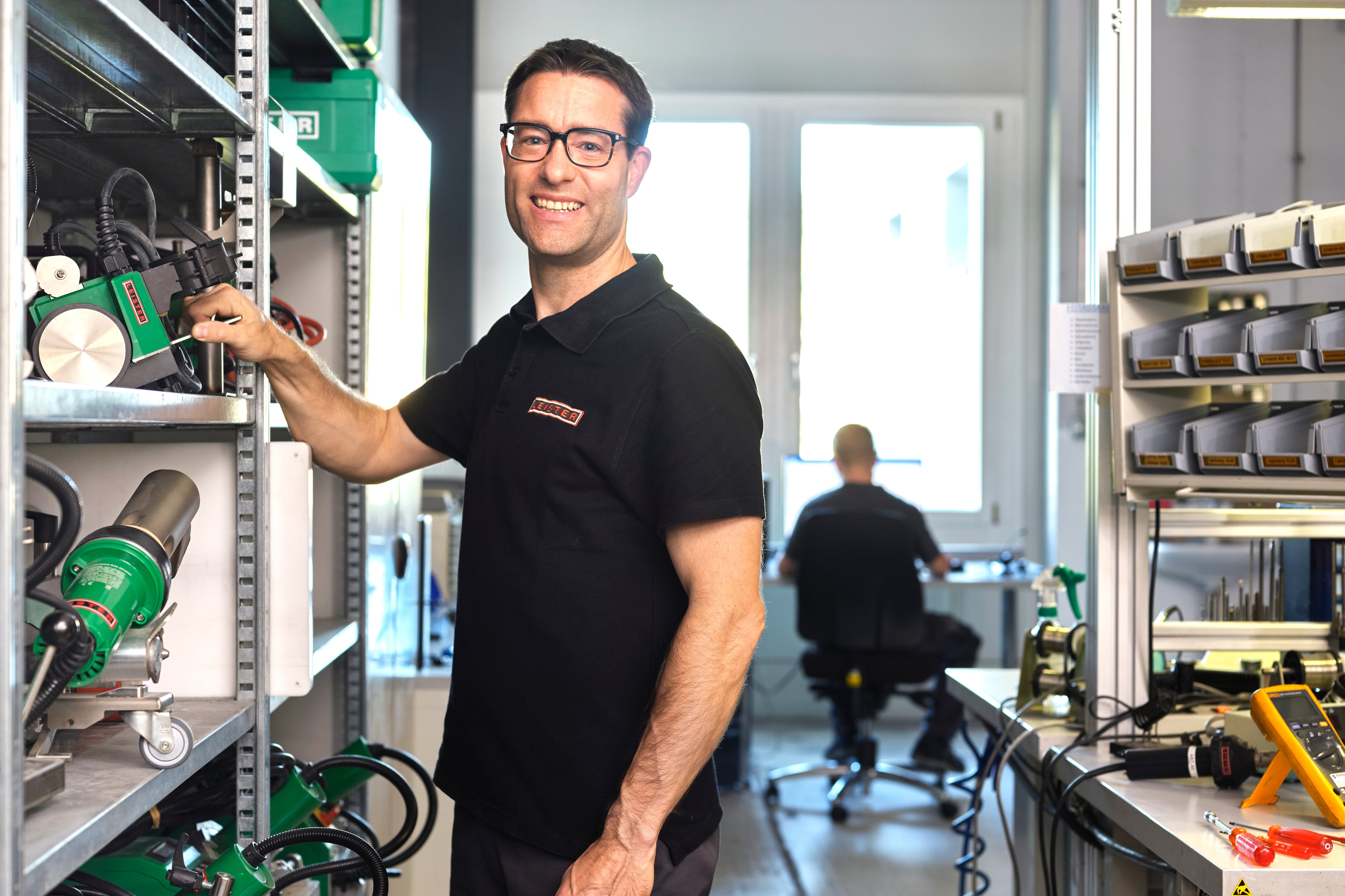 Técnico en uniforme negro trabaja en un dispositivo electrónico en una estación de trabajo en un laboratorio técnico bien iluminado, con herramientas y paneles de control visibles en el fondo.